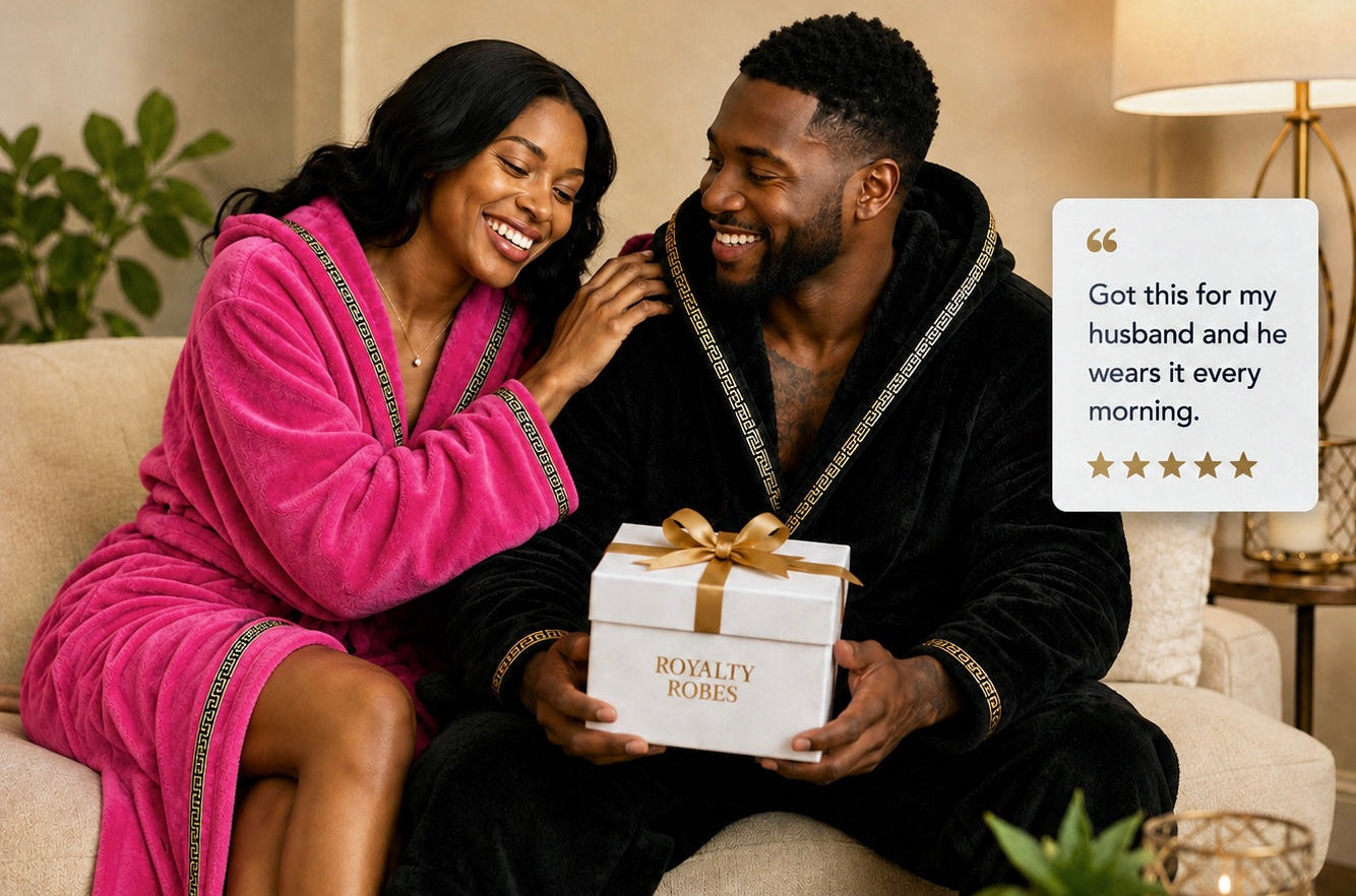 A couple sits on a beige sofa in a warmly lit living room, smiling at each other while the man holds a white gift box tied with a gold ribbon. The woman wears a bright pink plush robe with dark trim, and the man wears a black plush robe with gold trim.