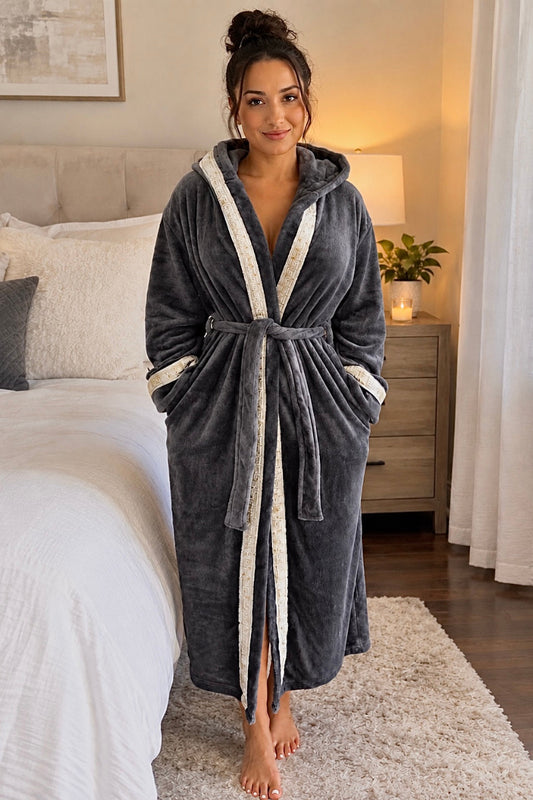 A woman wearing a plush dark gray hooded robe with silver and gold Greek key trim stands barefoot in a bedroom next to a bed.